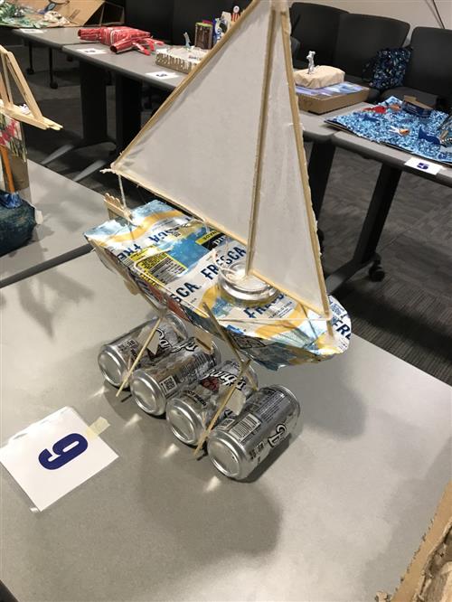 Student created boat made of recycled materials such as blue and yellow, cans, paper, and sticks.  